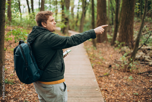 Blonde young 30s man wearing backpack holding phone and pointing something on city autumn park street. Urban lifestyle, travel concept. High quality photo
