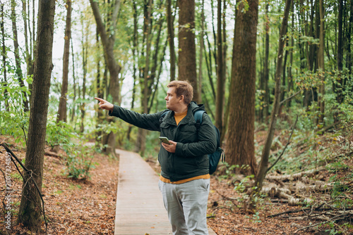 Blonde young 30s man wearing backpack holding phone and pointing something on city autumn park street. Urban lifestyle, travel concept. High quality photo