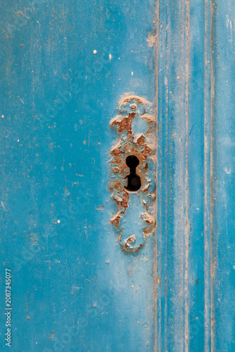 A rusty ornate keyhole escutcheon on a weathered blue-painted wooden door, showing peeling paint and aged patina