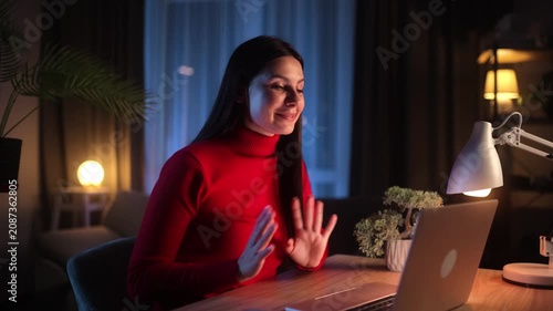 A young brunette woman in a red turtleneck sits at a wooden desk at night, smiling and gesturing with open hands during a video call or online meeting via her laptop in a cozy home office