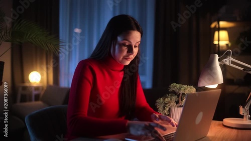 A tired young brunette woman in a red turtleneck touches her forehead, looking exhausted while working on her laptop late at night in a cozy home office, suffering from a headache.