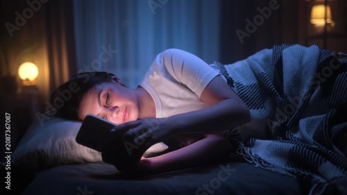 A tired young brunette woman lies in bed at night, staring at her smartphone screen and scrolling through social media while suffering from insomnia and sleeplessness.