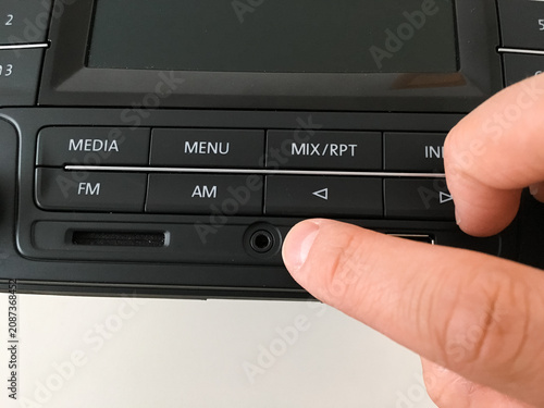 Finger pressing a button on a car radio faceplate with media menu and FM AM controls.