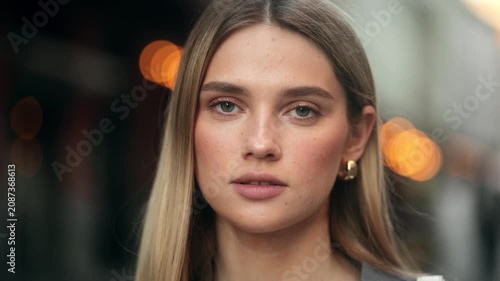 Close-up portrait of a serious young blonde woman looking directly at the camera on a city street at dusk with warm blurry bokeh lights in the background.