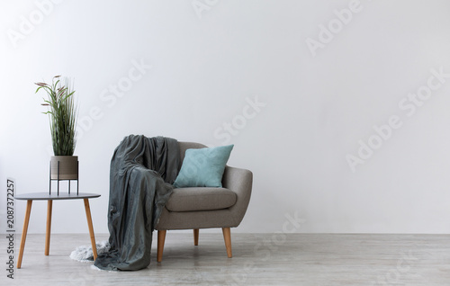 Modern boho cozy corner of home in living room. Gray vintage armchair with blue pillow and blanket, table with green plant in pot, wooden floor, on light wall background, copy space, mockup, nobody