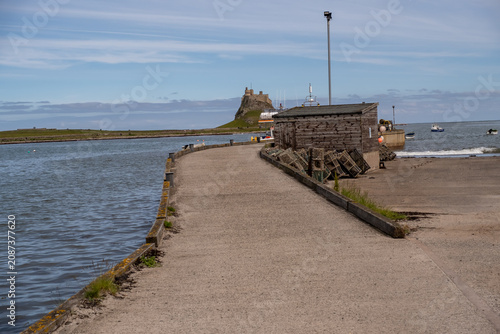 A pier on Holy Island, Northumberland coast, stretching into the sea as waves lap against its structure, framed by the island’s rugged and atmospheric shoreline.