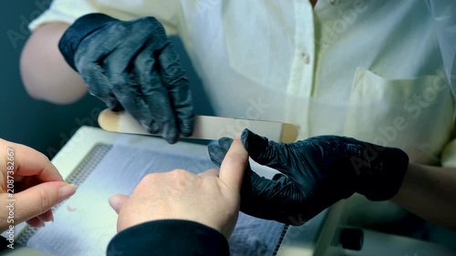 Close up professional manicure by female nail technician wearing glasses yellow shirt and black latex gloves providing careful nail treatment for woman in modern beauty salon