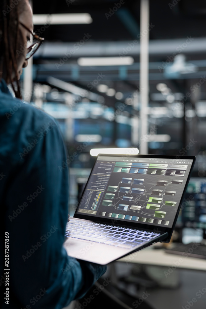 Naklejka premium Data center administrator uses a laptop to investigate neural network configurations, working with AI technology and high performance systems.