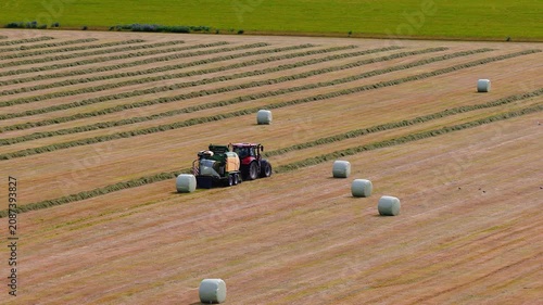 Aerial view of southern Iceland farmland near Skogafoss. A red tractor tows a baler, forming and wrapping silage bales on windrowed stubble under soft natural light.