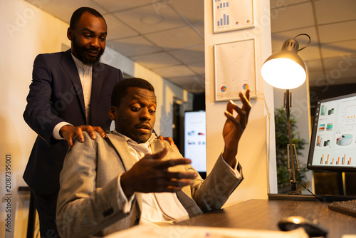 Vászonkép Black man taps tired colleague on shoulder at computer desk to stop him falling asleep, showing sleep deprivation during late night office work