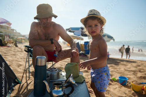 Tableau sur toile Father and toddler spend a sunny beach day together with sun hats, kettle and toys on sand near the sea, capturing family bonding, summer relaxation and playful childhood moments