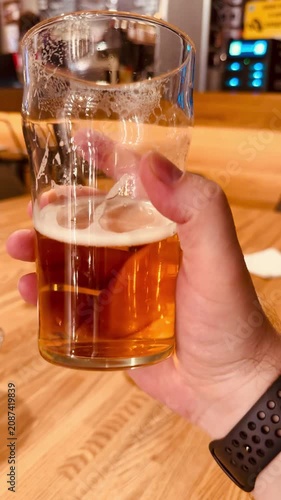 A man's hand holds a half-full glass of light beer above a wooden table in a beer pub. The setting is casual and inviting, with a blurred background