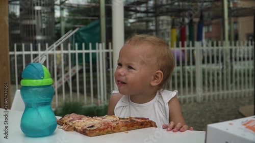 A baby girl with short blond hair takes a bite out huge pizza on the table, chews, picks corn with her fingers puts mouth. She chews appetizingly, squints sun, looks around.