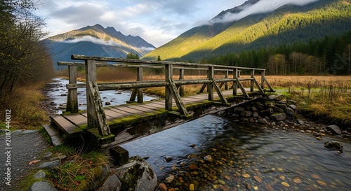 Rustic wooden bridge spans calm river in sunlit mountain landscape