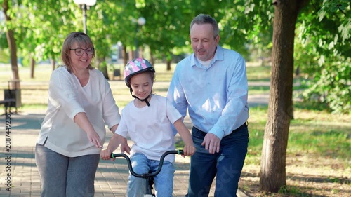 Dad mom teach daughter ride bike in park in summer. Father, mother teach child to maintain balance while riding bike. Family, parents, child walk together in park. Girl dream ride bike. Family sports