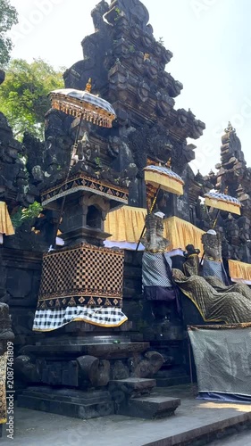 Ceremonial temple facade with draped guardian statues, fringed umbrellas, carved stone details, and checked textile accents under bright midday sun, with temple, ceremony, statue, guardian detail