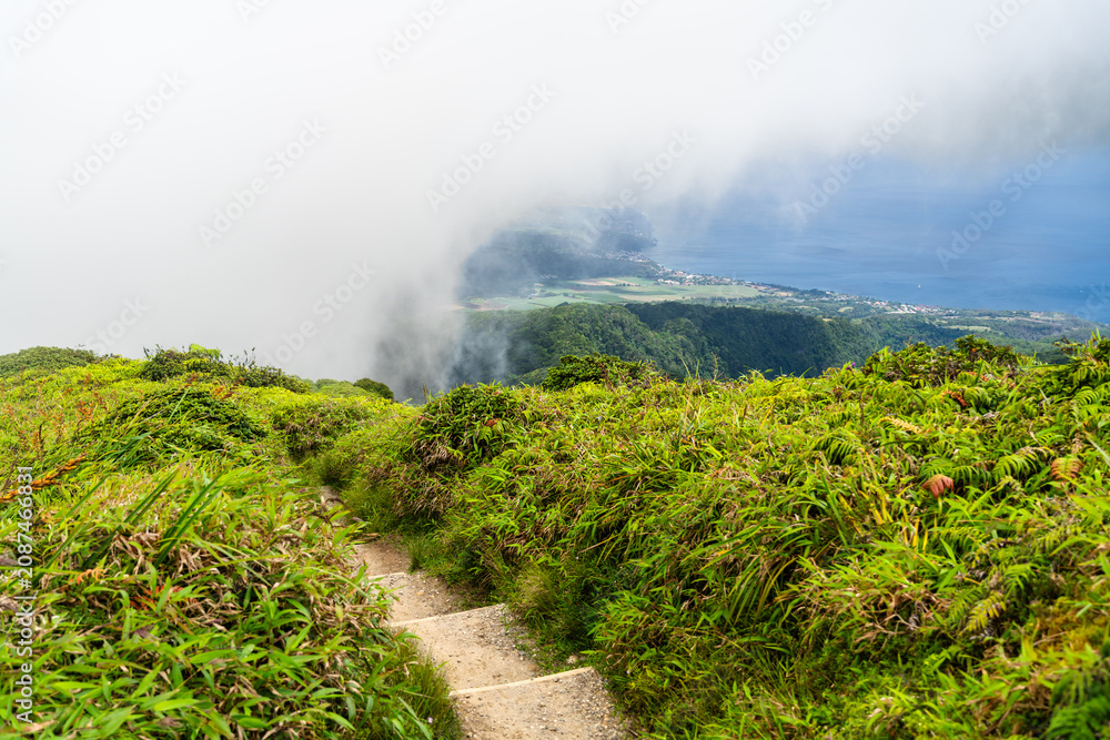 Naklejka premium Hiking path leading up Mount Pelee in Martinique, showing a scenic mountain trail with green grass, rolling clouds, and the ocean below