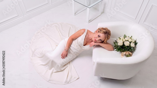 A bride in a white satin gown lounges on a white marble floor, resting against a modern armchair beside a white rose bouquet. Minimalist composition.