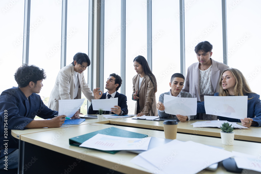 Fototapeta premium Diverse group of young professionals sitting at large conference table holding documents feeling focused and engaged during important modern corporate team meeting in bright glass walled office