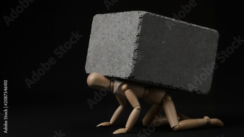 Wooden figure bears heavy concrete block, symbolizing burdensome weight.