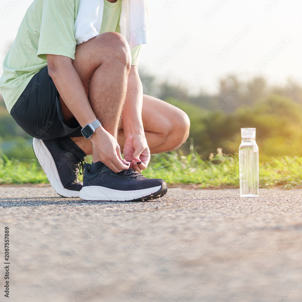Obraz premium Asian man with towel tying running shoes in the park with a reusable water bottle. male runner getting ready for jogging and fitness exercise. Healthy lifestyle and sustainability concept