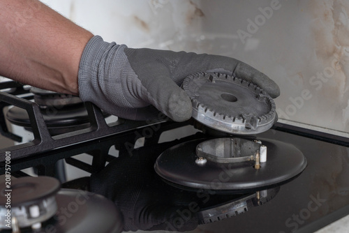 Close-up of a worker in a protective glove holding and placing a metal burner crown diffuser onto a black glass gas hob during appliance service, assembly, or kitchen maintenance.