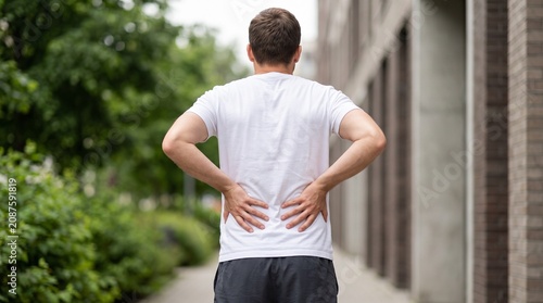 Man suffering back pain standing outside in a white t-shirt while holding his lower lumbar area due to a severe muscular injury during outdoor exercise