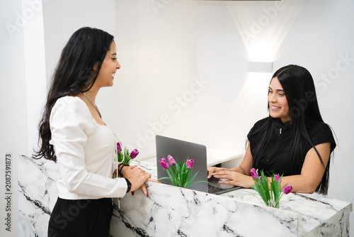 Foto Two young adult Hispanic women communicating at a modern marble reception desk with a laptop