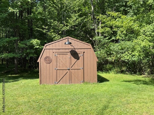 Obraz na plátně Brown wooden backyard storage shed with double barn style doors and exterior gooseneck light surrounded by green lawn and mature forest in the Midwestern United States
