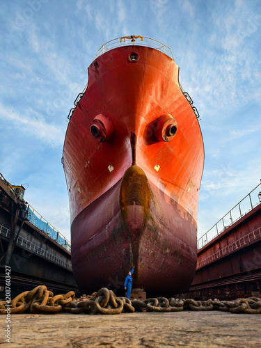 A shipyard worker stands proudly in front of a giant cargo ship under maintenance in a dry dock.