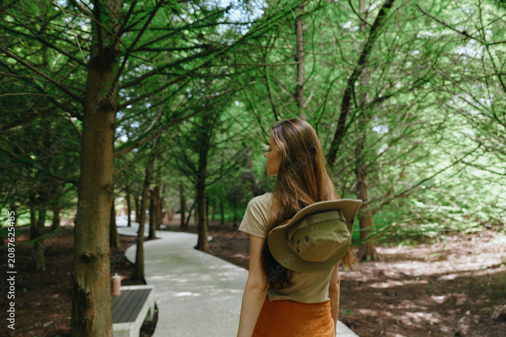 Naklejka premium Woman walking on forest trail with backpack and hat, back view on shaded park path surrounded by green trees and nature, dappled sunlight and casual summer outdoor stroll.