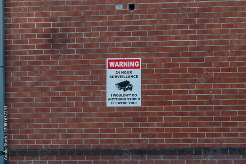 фотографія Humorous 'WARNING 24 HOUR SURVEILLANCE' sign on a red brick wall with a security camera icon, playfully stating 'I WOULDN'T DO ANYTHING STUPID IF I WERE YOU!' – a deterrent for mischief