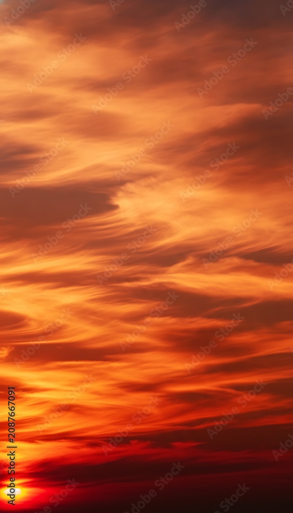 Fototapeta premium Intense orange and red sunset sky with swirling, wispy cloud formations creating a fiery and dramatic atmosphere as the sun dips below the horizon, casting a warm glow across the wallpaper background