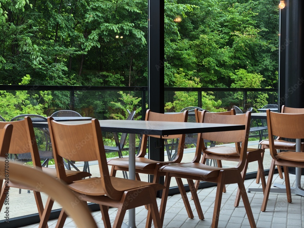 Fototapeta premium Empty chairs line a modern cafe patio. Large windows frame lush green forest beyond. Natural light bathes the scene in calm tranquility. Wooden tables and chairs invite quiet conversation