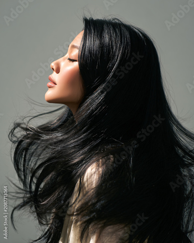 Beautiful Asian woman with long healthy glossy black hair flipping her hair in a luxurious salon, dramatic studio lighting mixed with natural window light, silky hair movement