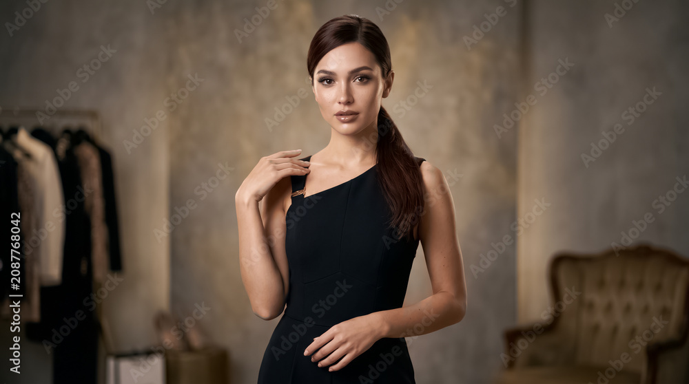 Fototapeta premium Beautiful young woman with dark hair wearing a stylish black dress poses elegantly for a professional high quality fashion photograph