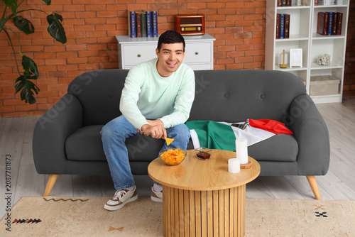 Young man with Mexican flag eating nachos with sauce at home