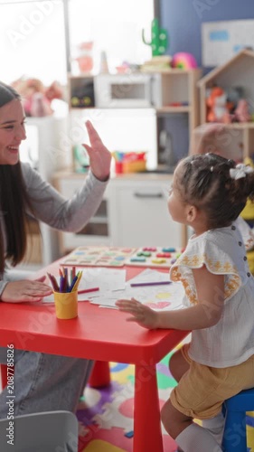 Woman gives high five with hand to little girl over drawings on red table in building; play learning bonding joy.
