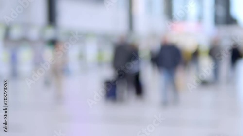 Blurred travelers move through bright, modern transit hall. People carry luggage, walk casually, and appear focused on journeys. Scene suggests airport or train station ambiance