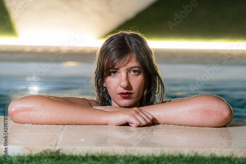 Young woman with wet hair leaning on the edge of an illuminated outdoor swimming pool at night, making direct eye contact with the camera. Dramatic lighting creates a cinematic atmosphere.