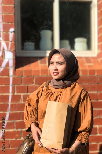 Stylish young Asian Muslim woman wearing a modern hijab and elegant casual outfit while holding a paper shopping bag outdoors, showcasing authentic modest fashion, contemporary personal style.