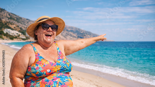 Joyful plus-size woman in a floral swimsuit pointing at the sea on a beach
