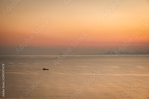 A small fishing boat sailing on the calm ocean with a distant industrial cityscape under sunrise sky.