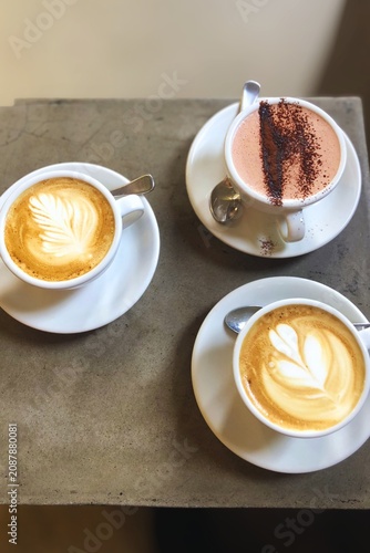 Top view of specialty coffee drinks served on a concrete café table. Beautiful latte art cappuccinos paired with a creamy hot chocolate topped with cocoa powder, creating a cozy coffeehouse atmosphere