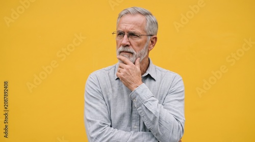 Elderly bearded man in glasses wearing blue shirt looking sideways with hand on chin on yellow background, thinking