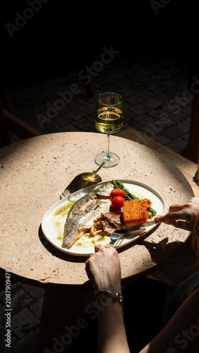 Slika na platnu Gourmet Meal by the Window: A delectable fish dish served with fresh vegetables and a glass of white wine, elegantly presented on a rustic table