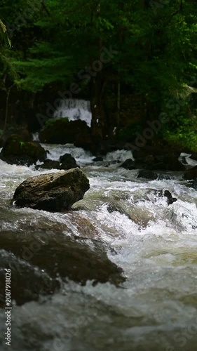 Vertical slow motion video of a powerful, rushing river stream cascading over rocks with a lush green jungle and a waterfall in the blurred background. 