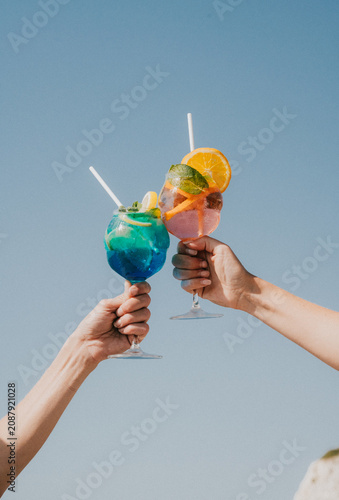 Photography Two hands clink glasses of colourful cocktails against a blue sky