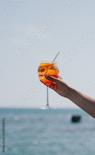 Billede på lærred Orange cocktail held aloft against a blue ocean backdrop during summer