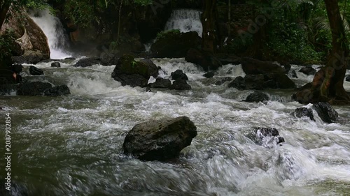 Powerful water rushing through dark rocks in a stream with beautiful waterfalls cascading in the background of a lush tropical rainforest. Thailand.